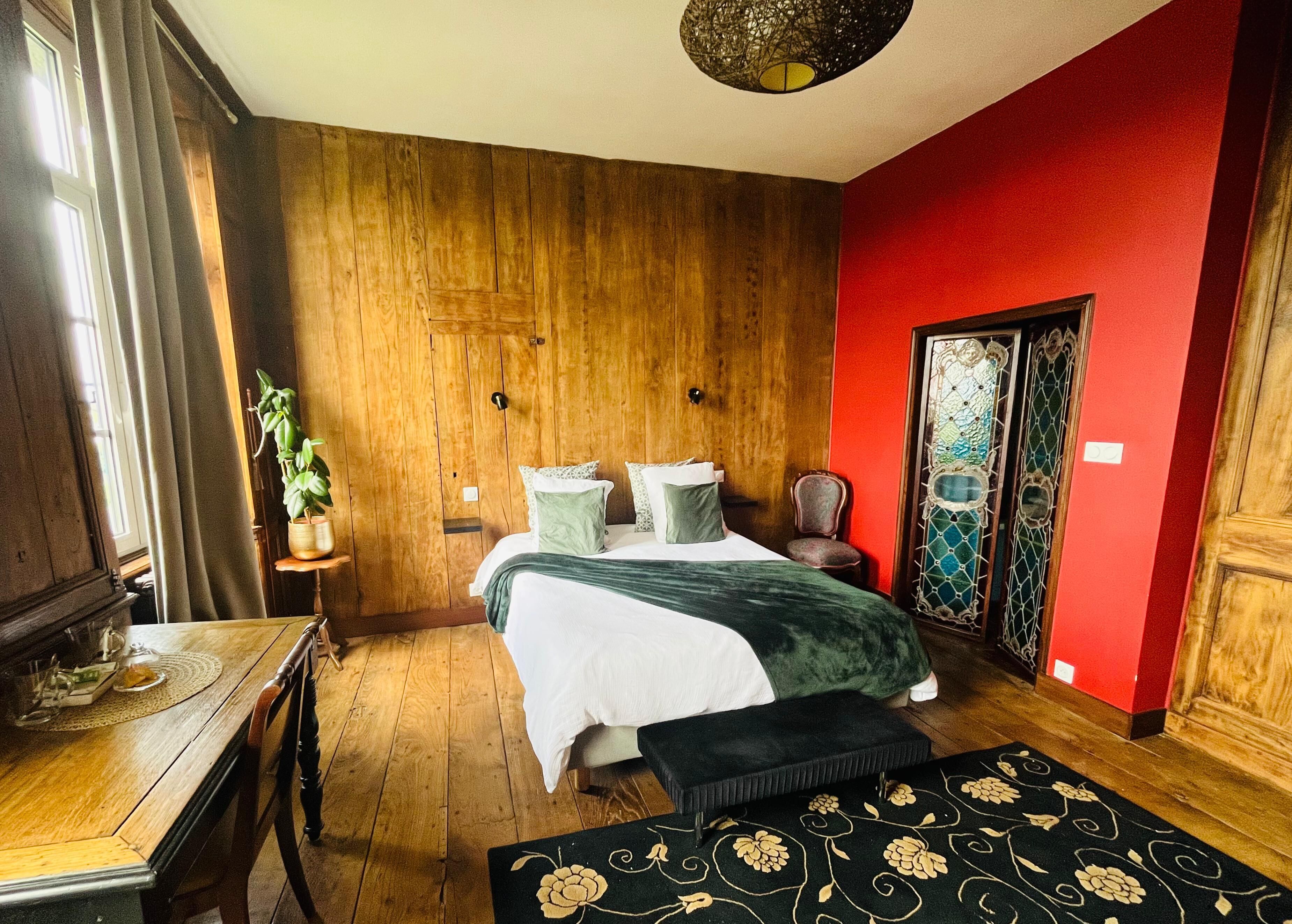 A bedroom with a wooden floor, a bed with a green blanket, a chair, a small table with a plant, and a red wall.
