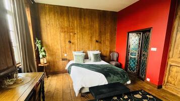 Un dormitorio con suelo de madera, una cama con manta verde, una silla, una mesita con una planta y una pared roja.