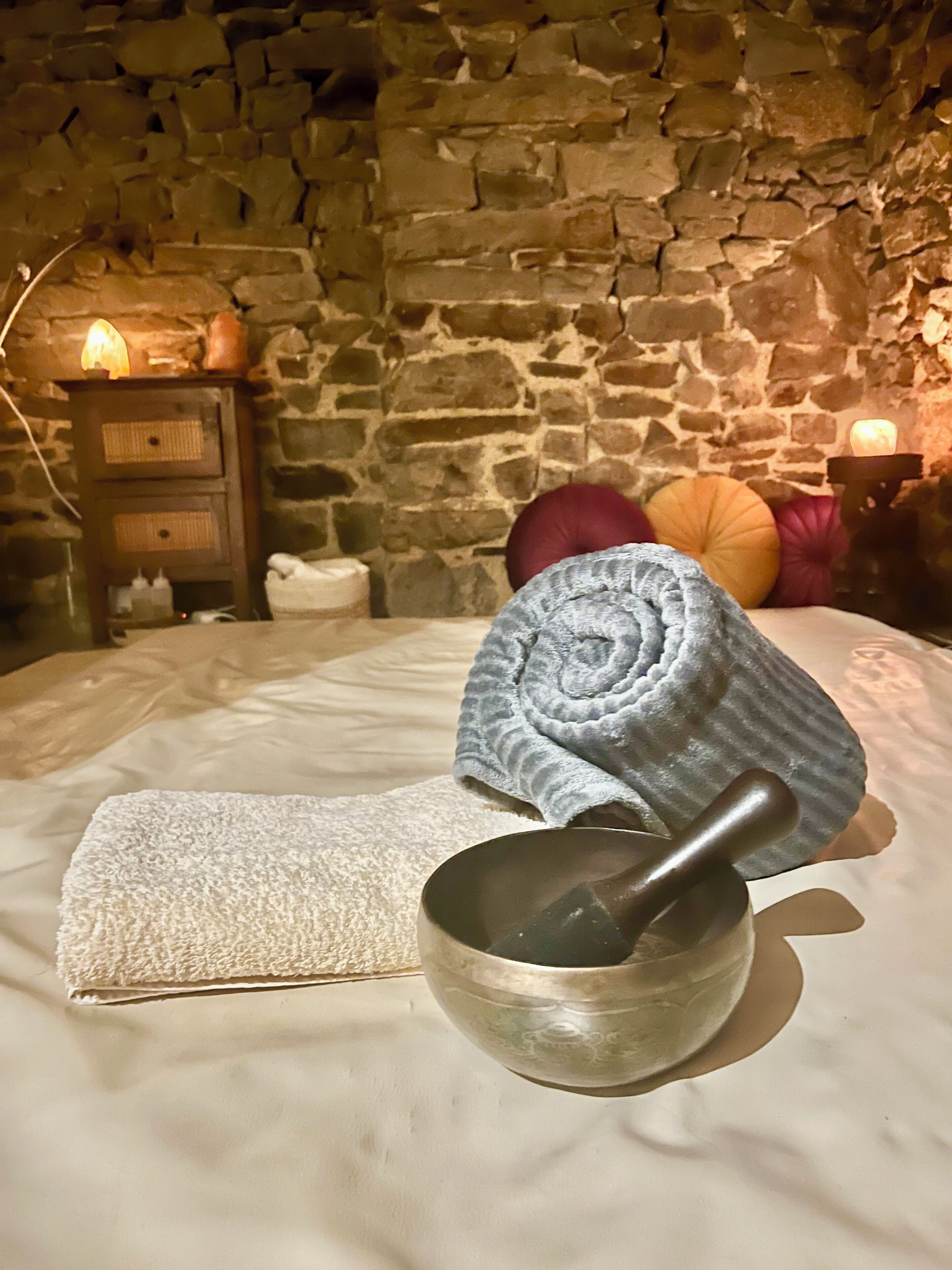 A stone-walled room with a bed, a wooden nightstand with a lamp, and a bowl with a pestle.