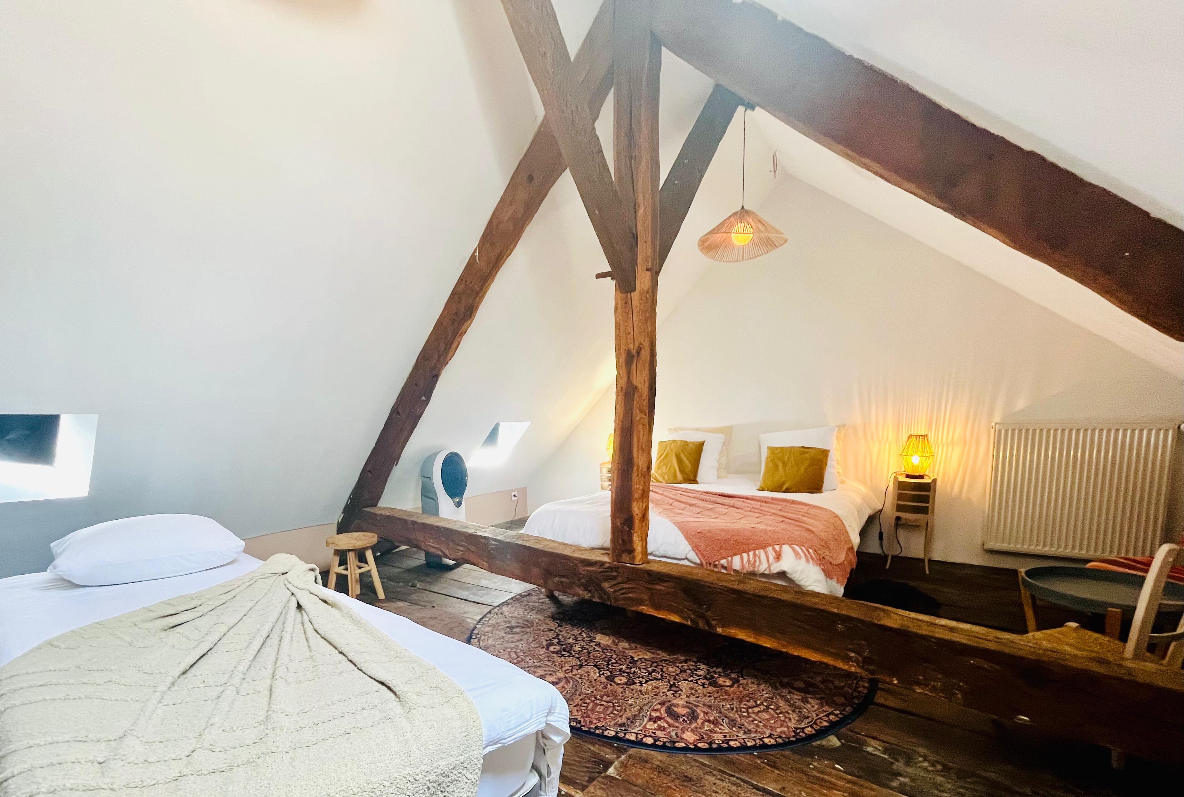 A room with a sloped ceiling, wooden beams, a bed with a white and beige blanket, a small stool, a round patterned rug, a table, and a chair.