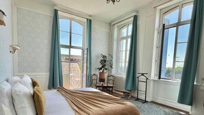 Une chambre à coucher comprenant un lit, une commode en bois, une petite table, une chaise, une plante en pot et une vue sur l’extérieur grâce à de grandes fenêtres.