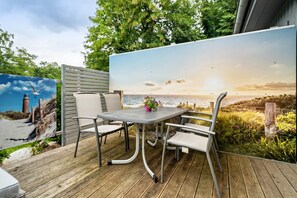 Una terraza de madera con una mesa y sillas de metal, una planta en maceta y un mural con atardecer sobre el mar.