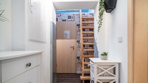 Ein schmaler Flur mit einer weißen Kommode, einer kleinen Pflanze auf einem weißen Beistelltisch und einer Holztür, die zu einem Raum führt, dessen Wand mit gerahmten Gegenständen gestaltet ist.