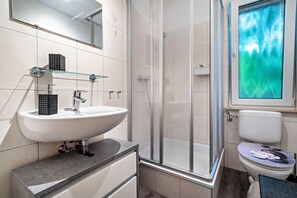 Un baño moderno con cabina de ducha de vidrio, un lavamanos blanco con dispensador de jabón negro y un inodoro con tapa decorativa.