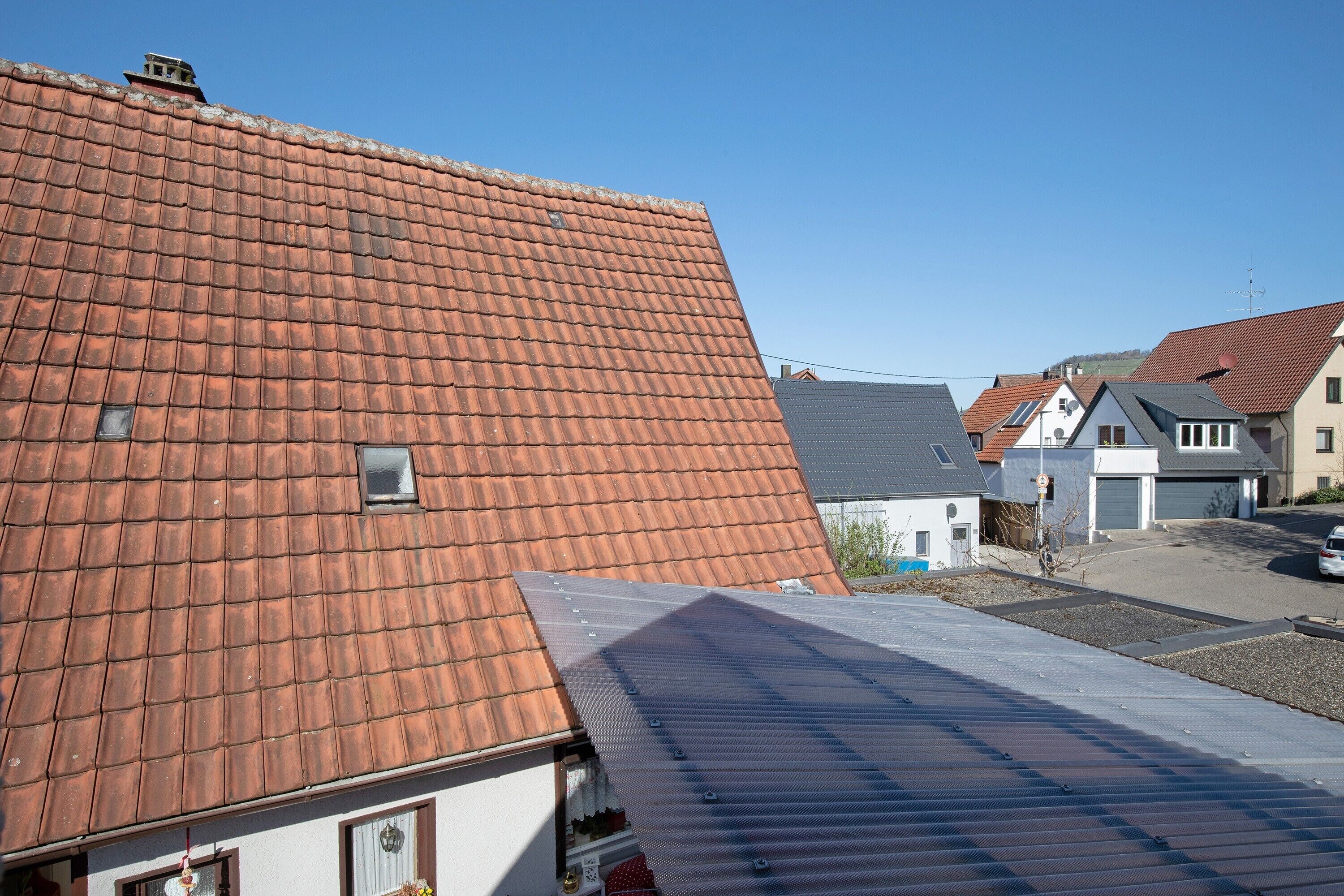 A residential area with a red-tiled roof, a grey metal roof, and a clear blue sky.