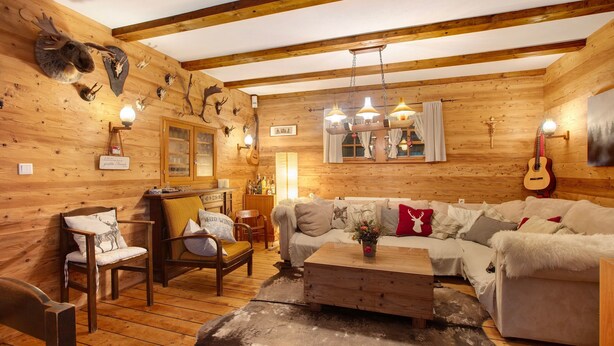 Interior de una cabaña de troncos con suelo de madera, un sofá, sillas, una mesa de centro y monturas de animales en las paredes.