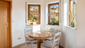 A corner room with a round wooden table, white chairs, and two windows with views of a brick wall and plants.