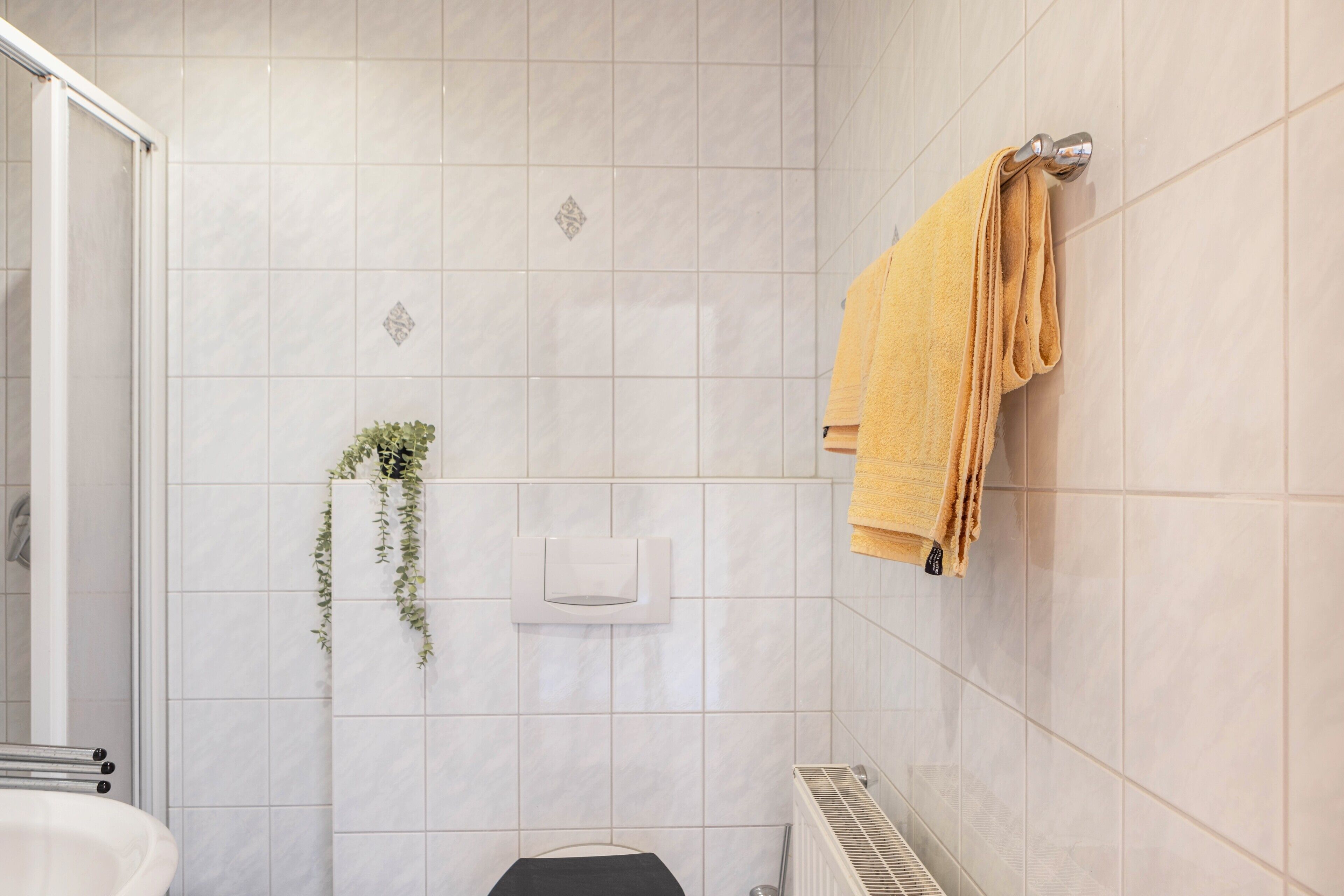 Een badkamer met witte tegels, een handdoekhouder met een gele handdoek, een plant en een toilet.