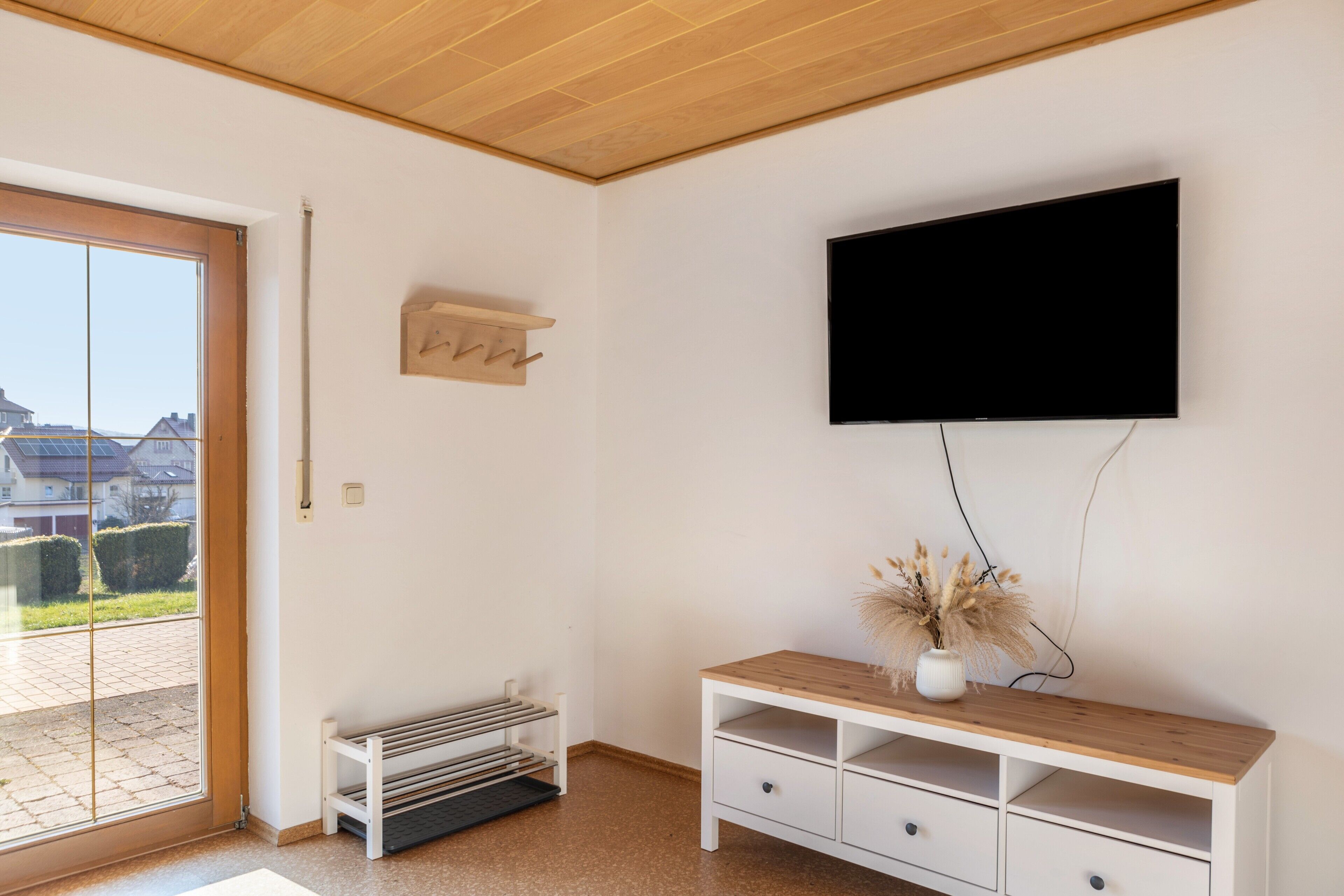 A room with a television mounted on the wall, a wooden console table with a vase, a bench with magazines, and a door leading outside.