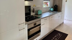 A modern kitchen with white cabinets, a gas stove, an oven, a sink, and a window with a view of the outdoors. - Furnished Chalet Sint Maartenszee (Sint Maartensvlotbrug)