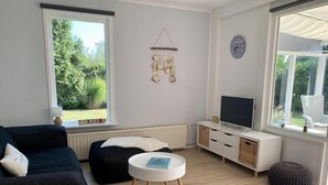 Ein Wohnzimmer mit einem dunklen Sofa, einem weißen Couchtisch, einem Fernsehgerät auf einer weißen Kommode und einem Fenster mit Blick auf Grünflächen.