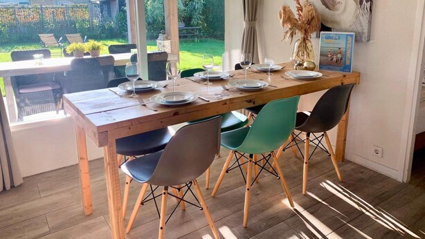 A dining area with a wooden table set for four, surrounded by modern chairs, with a view of a garden and a beach scene on the wall. - Home in Sint Maartenszee With Veranda (Sint Maartensvlotbrug)