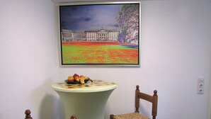 A room with a table and two chairs, a painting of a building on the wall, and a power outlet on the wall.