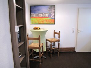 Ein Zimmer mit zwei Holzstühlen, einem kleinen Tisch mit Obst, einem Bild eines Gebäudes und einer Tür.