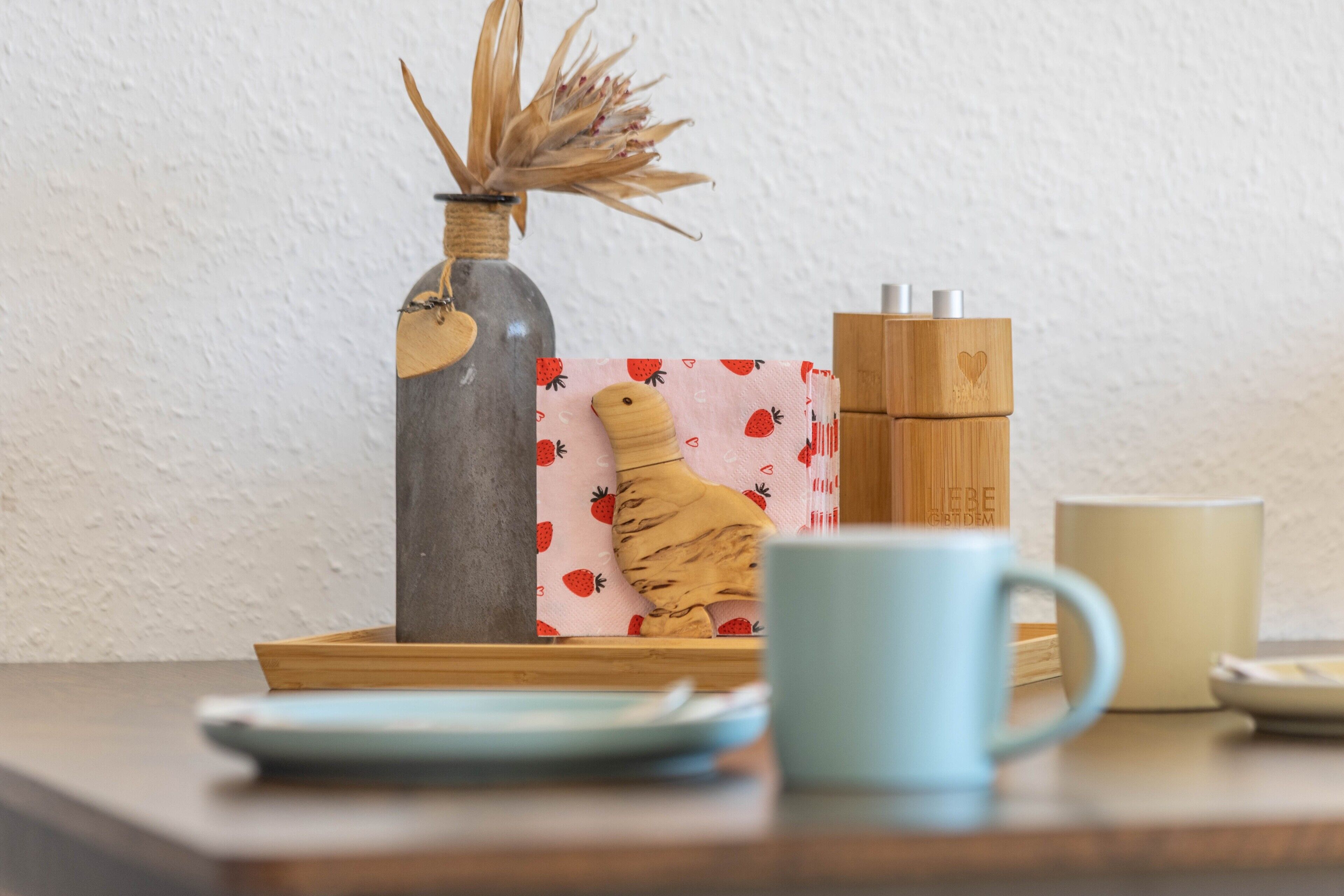 Una mesa con una taza azul, una planta pequeña en una botella, un bloque de madera con diseño de corazón y una caja de pañuelos rosa.