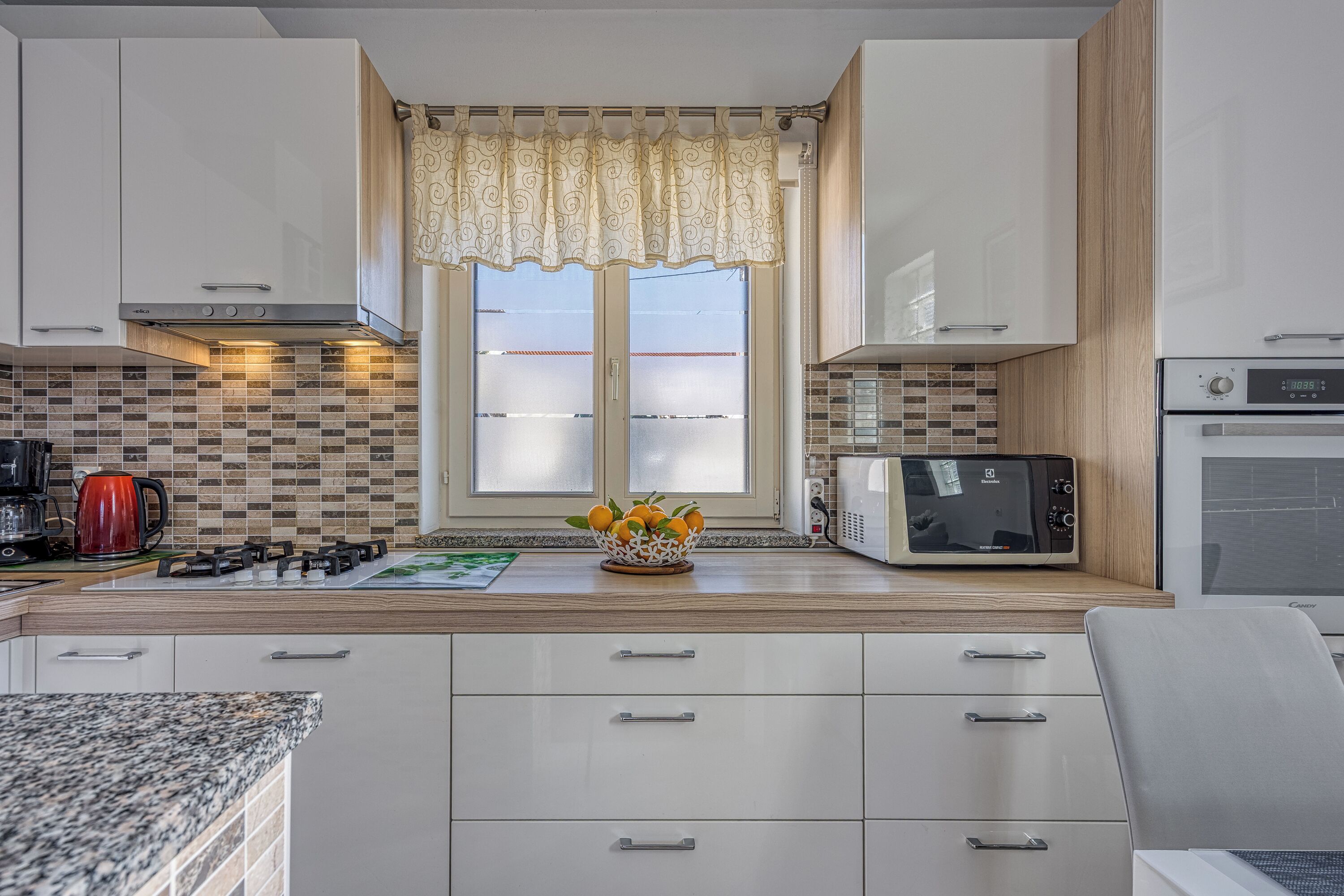 A modern kitchen with white cabinets, a gas stove, a microwave, and a window with patterned curtains.