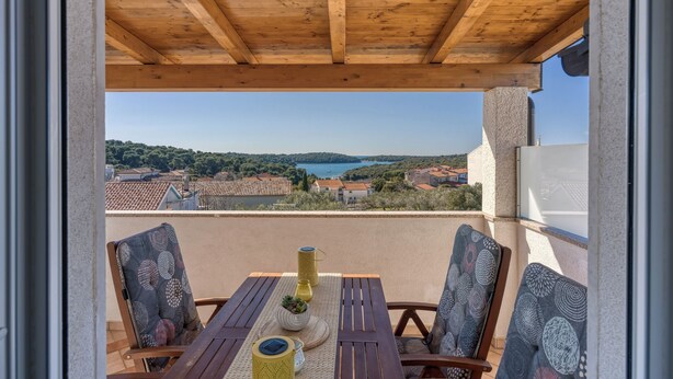 Un balcón de madera con una mesa y sillas, con vistas panorámicas a un lago y colinas circundantes.