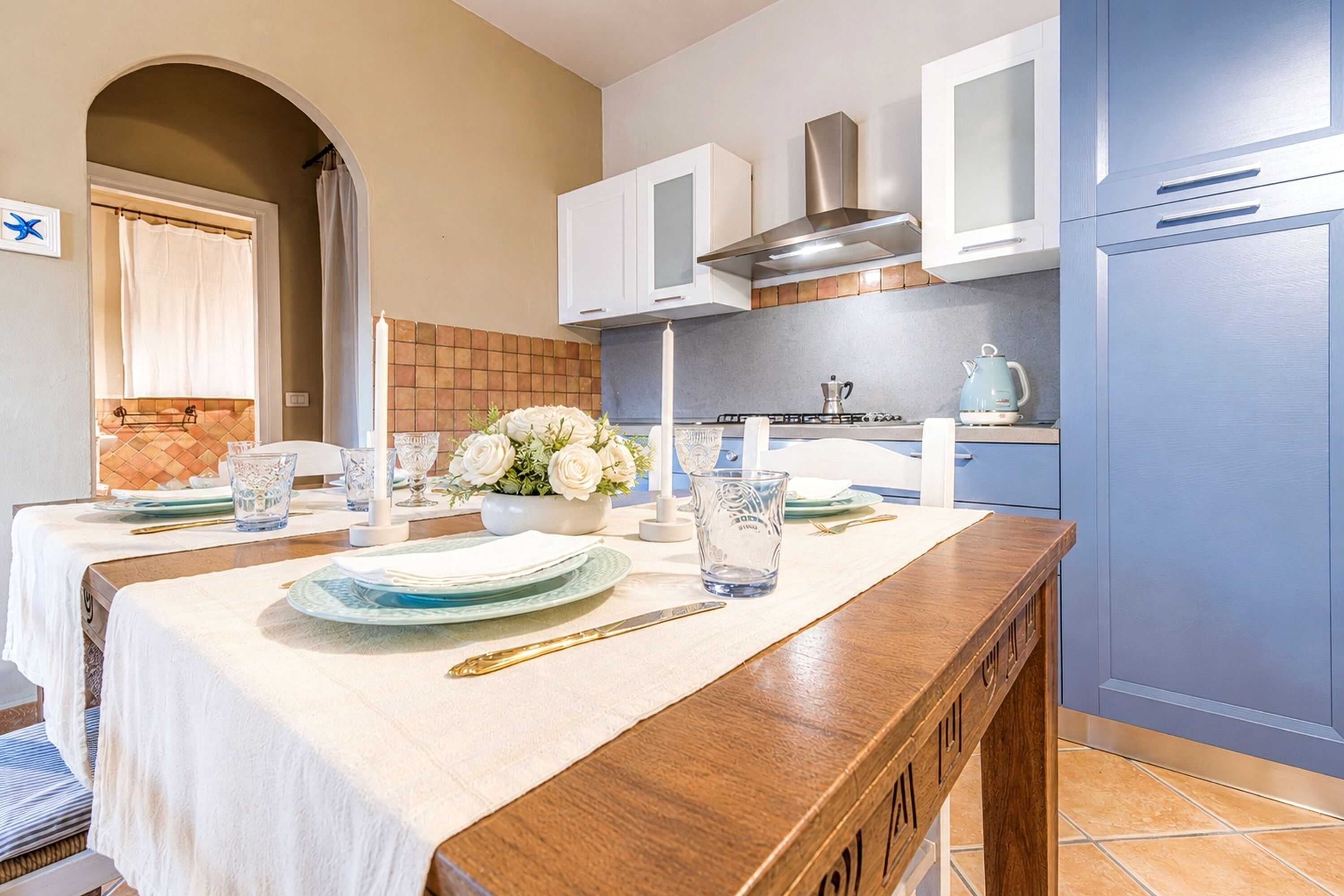 Cozinha com mesa de jantar posta para dois, armários brancos, coifa em aço inoxidável e geladeira azul.
