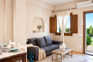 A living room with a sofa, a table, a chair, a window with curtains, and a view of greenery outside. - Dimora Apartments (Arzachena)