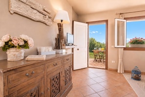A room with a wooden cabinet, a television, a door leading to an outdoor patio, and a window with a view of greenery.