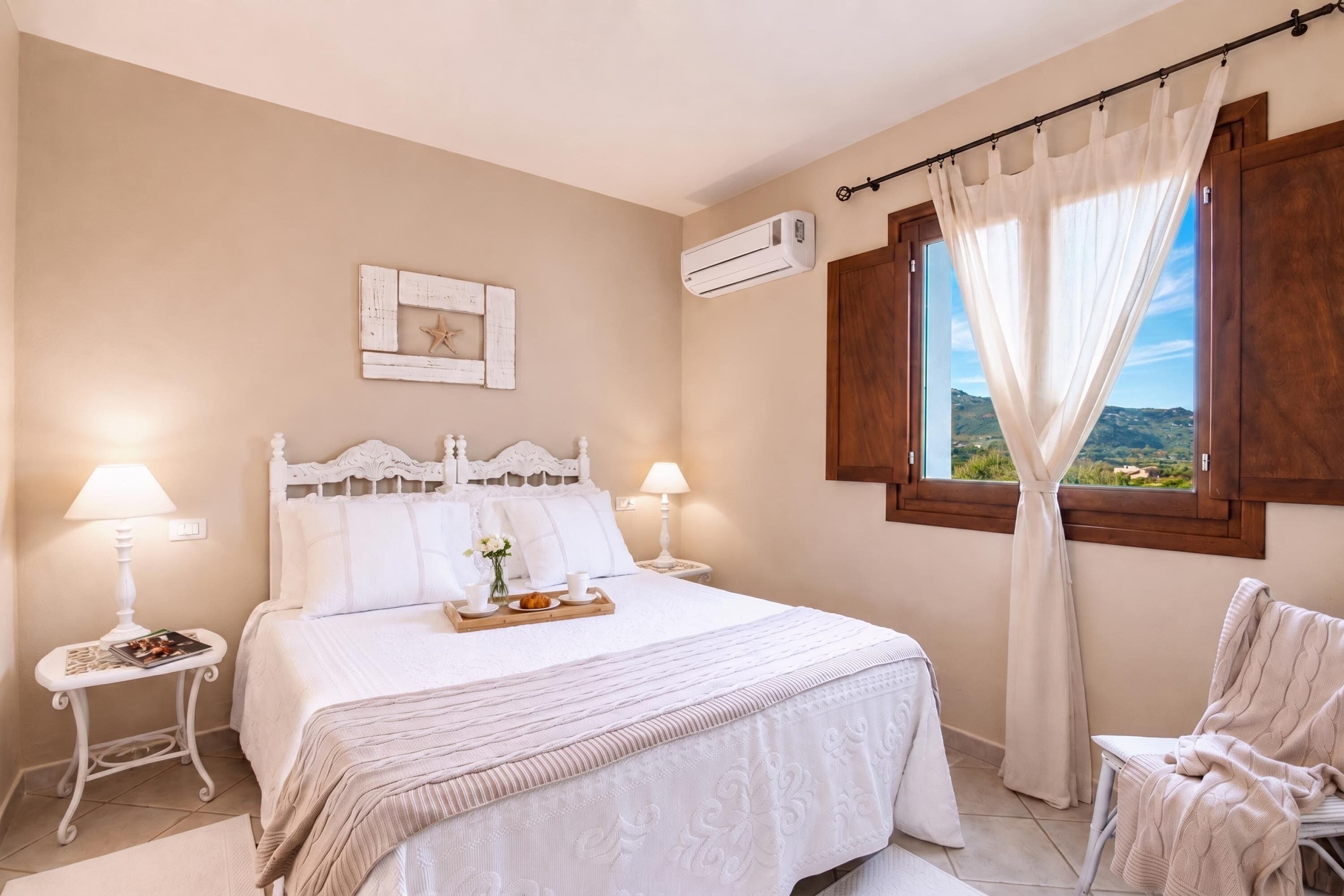 A bedroom with a bed, bedside tables, an air conditioning unit, a window with curtains, and a view of the landscape.