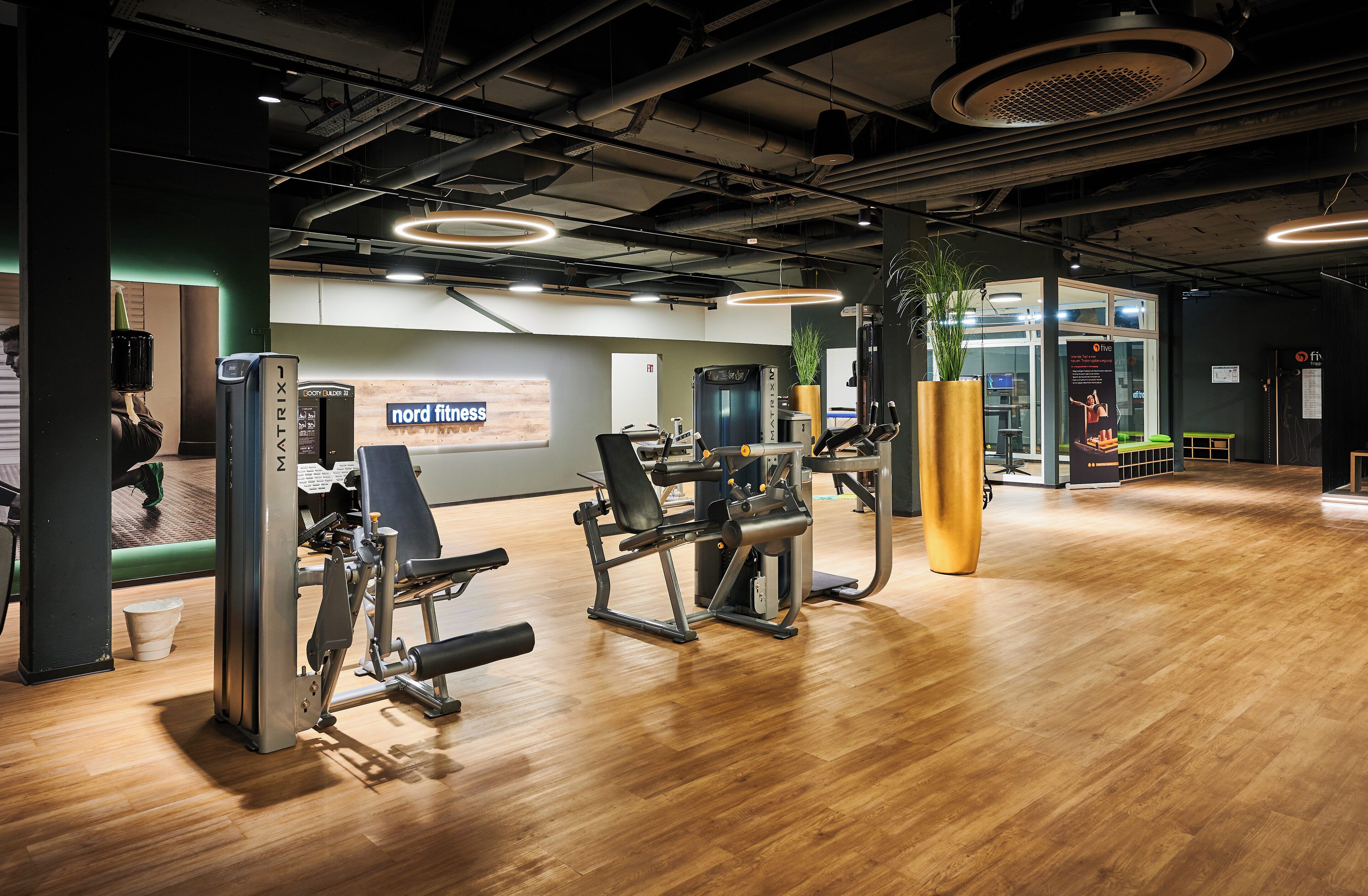 Ein Fitnessstudio mit verschiedenen Trainingsgeräten, Holzboden und einem Schild mit der Aufschrift 'Nord Fitness'.