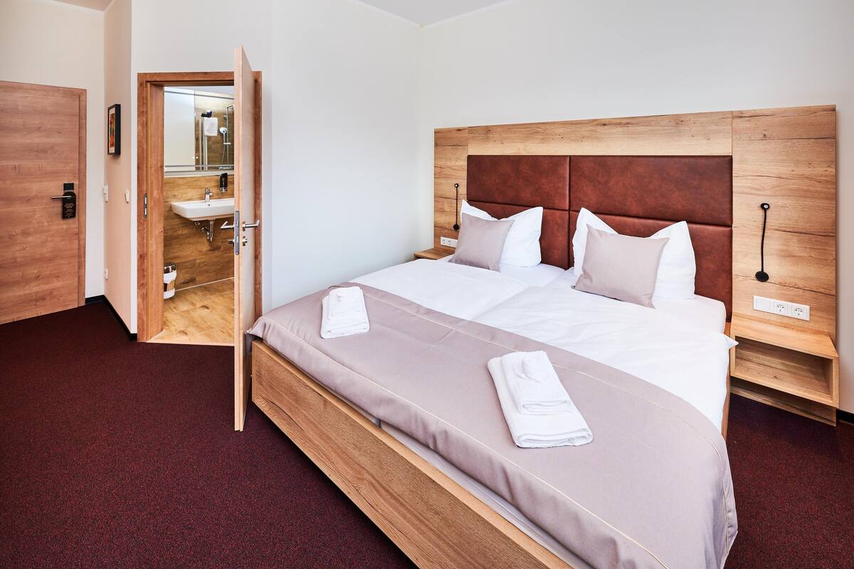 Una habitación de hotel moderna con cabecera de madera, una cama grande con colchón color beige, dos almohadas y una mesita de noche de madera.