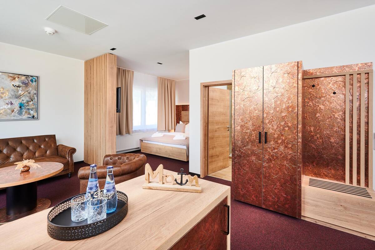 Una habitación de hotel moderna con un sofá de cuero marrón, una mesa de centro de madera con botellas de agua, una cama con cabecero de madera y un amplio armario de madera con detalles de cobre.