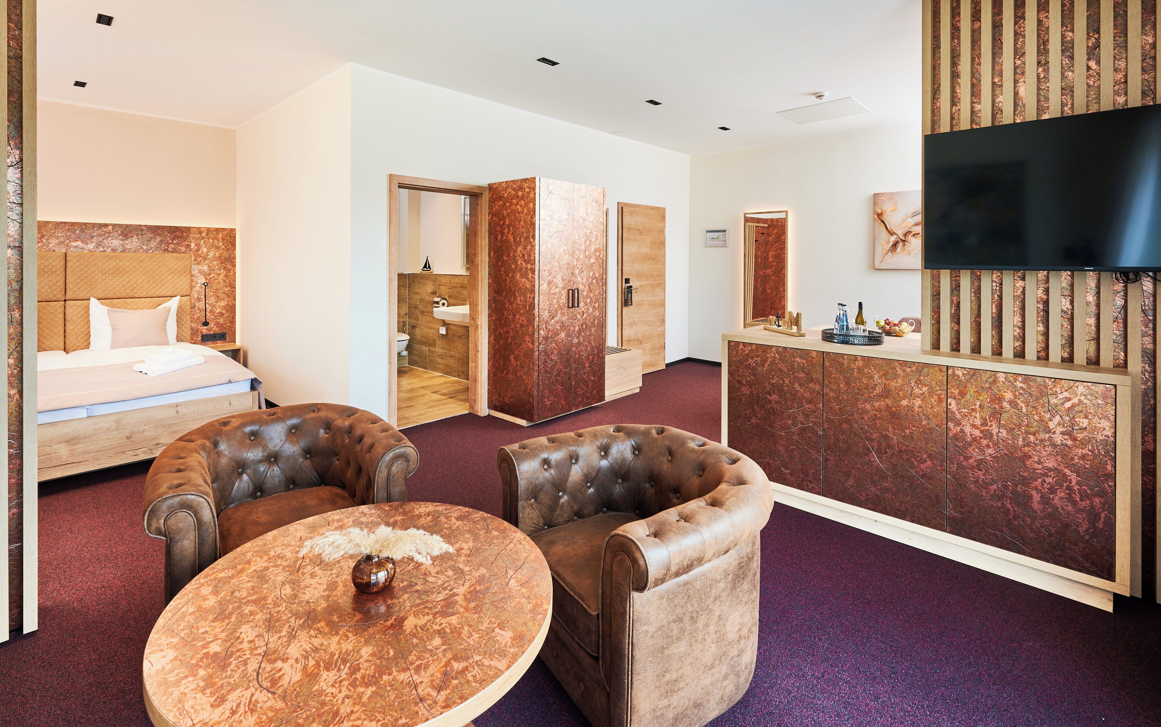 Habitación de hotel con cama, dos sillones, una mesa redonda de centro, televisión y baño visible a través de una puerta abierta.