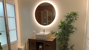 A bathroom with a wooden vanity, a round mirror with a light, and a potted plant.