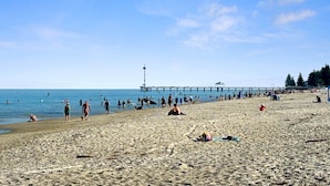 Ein Strand mit einem Steg, der sich ins Wasser erstreckt, Menschen schlendern und entspannen auf dem Sand, und ein klarer Himmel.