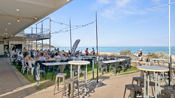 Ein Sitzbereich im Freien mit Tischen und Stühlen mit Blick auf einen Strand und das Meer.