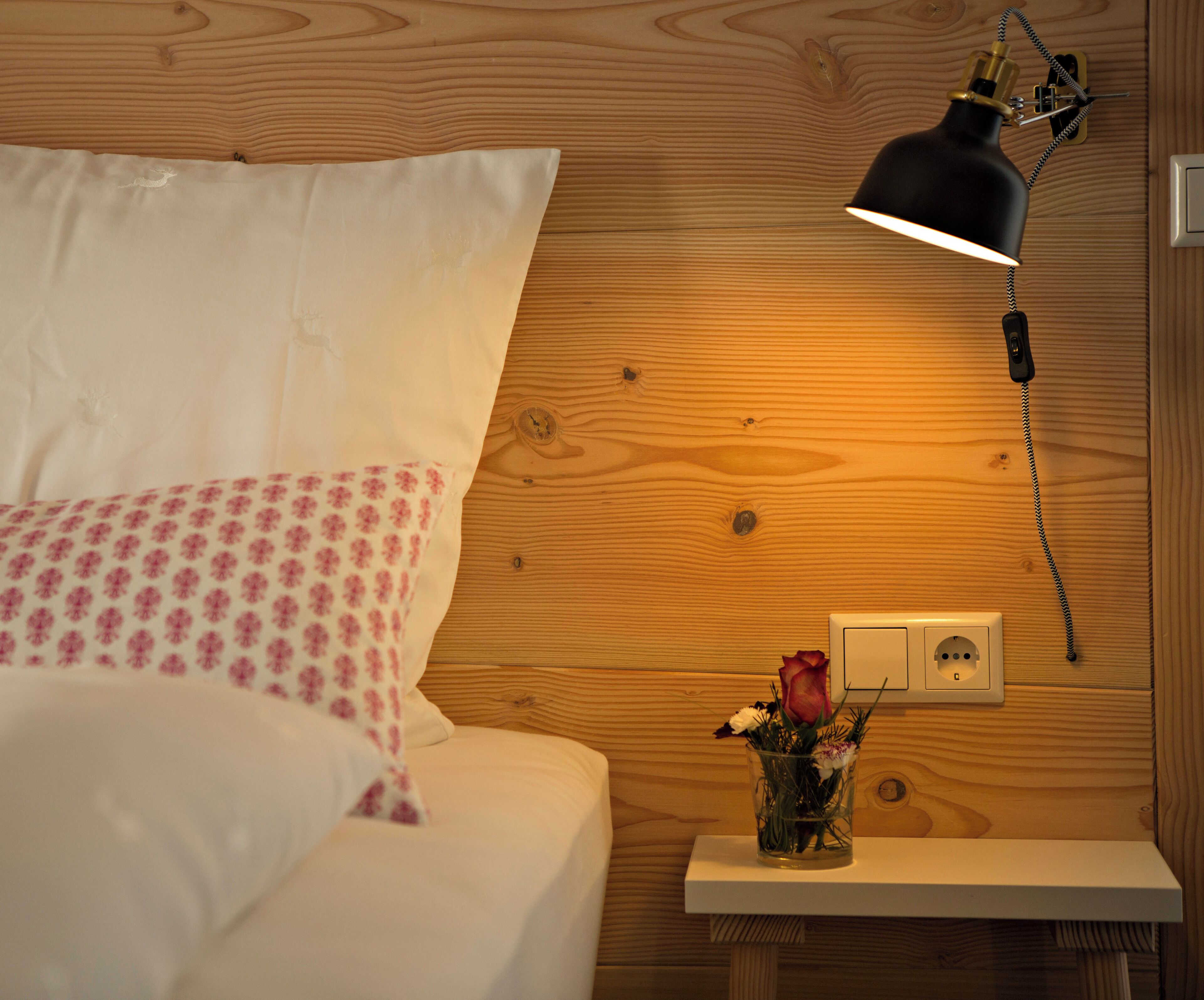 A neatly made bed with white and pink bedding, a bedside table with a vase of flowers, and a wall-mounted lamp.