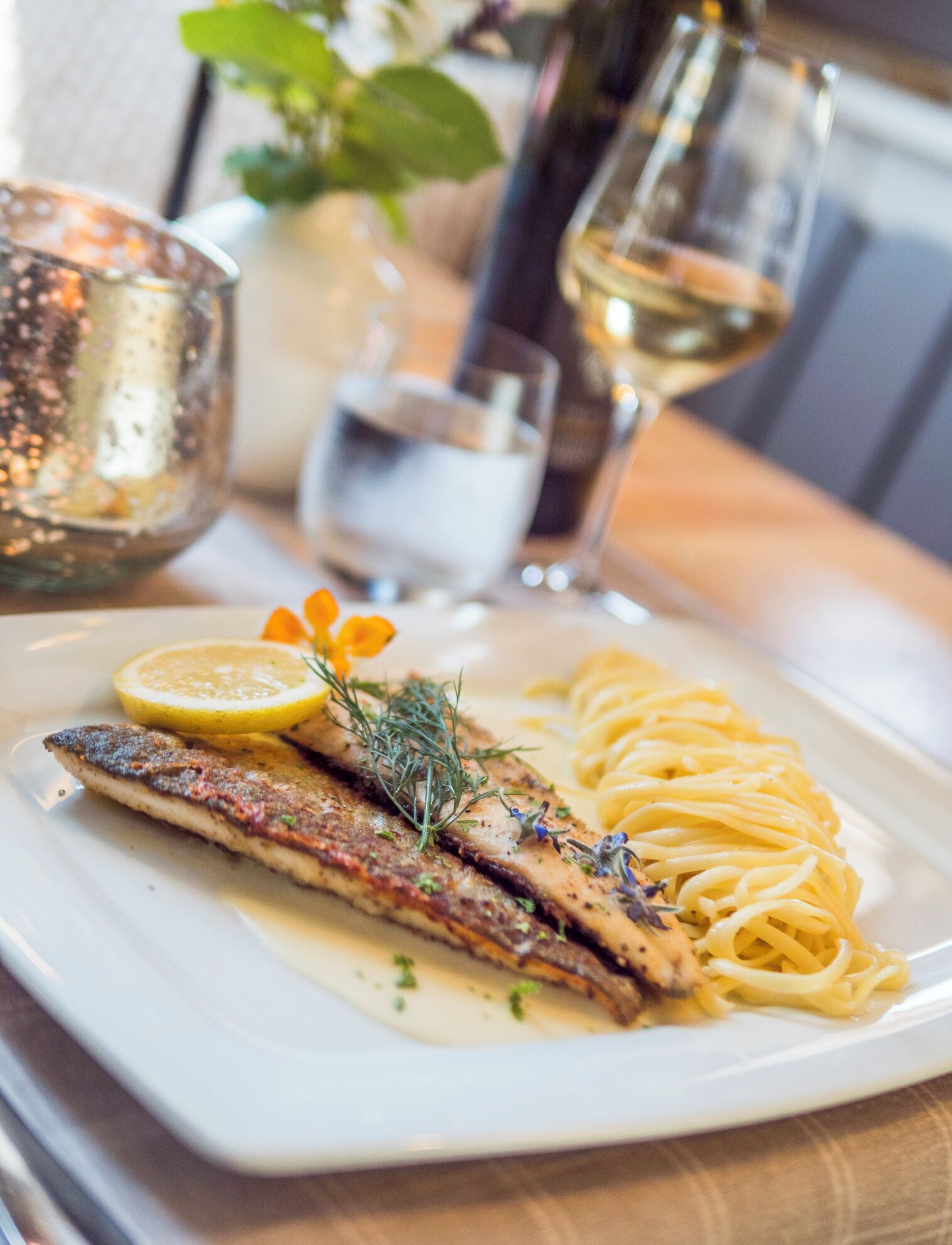 A plate of food with a fish fillet and pasta, accompanied by a glass of white wine and a candle.