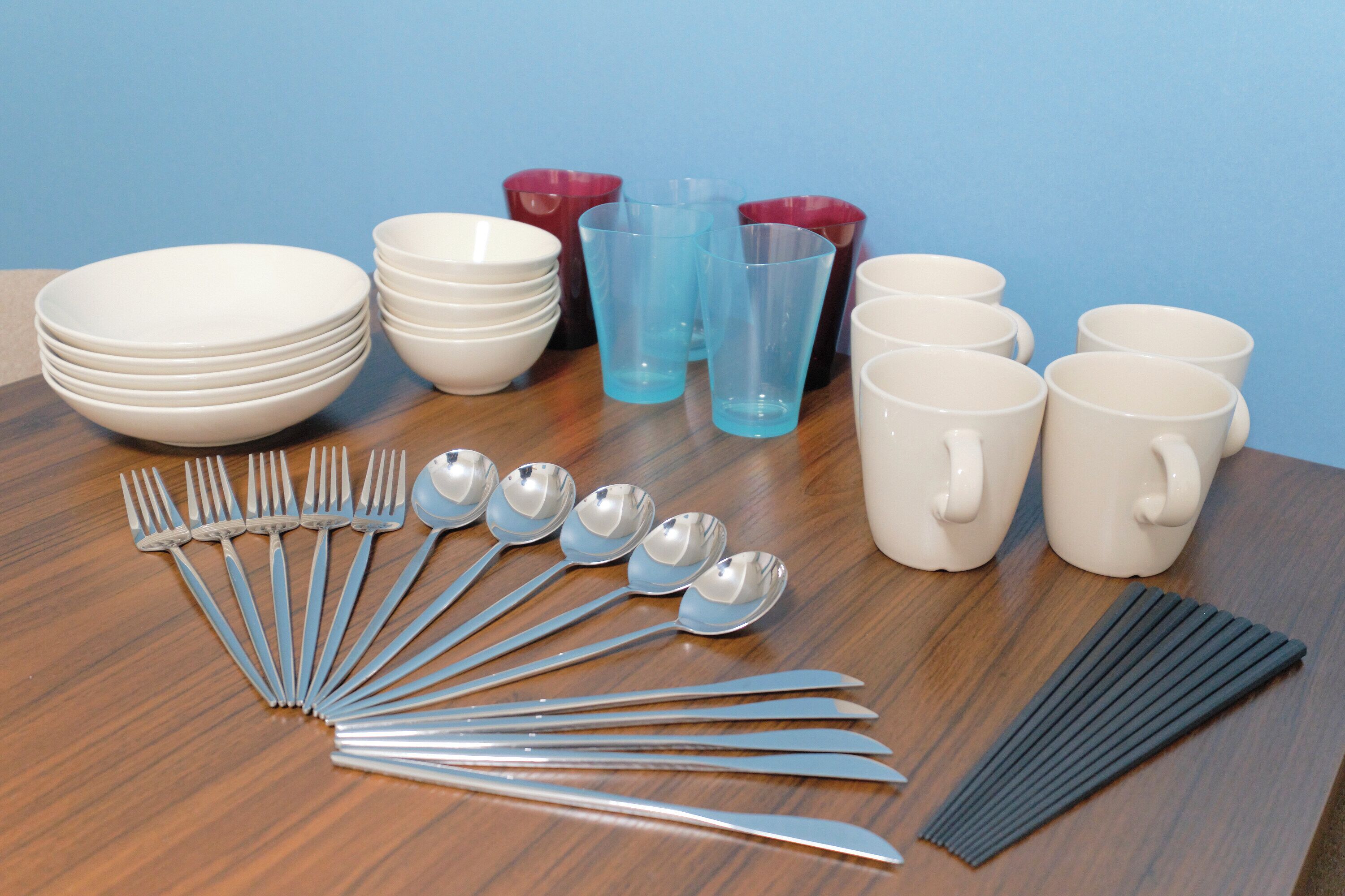 Una mesa con cuencos blancos apilados, vasos de plástico azul y rojo, tazas blancas y un juego de cubiertos.