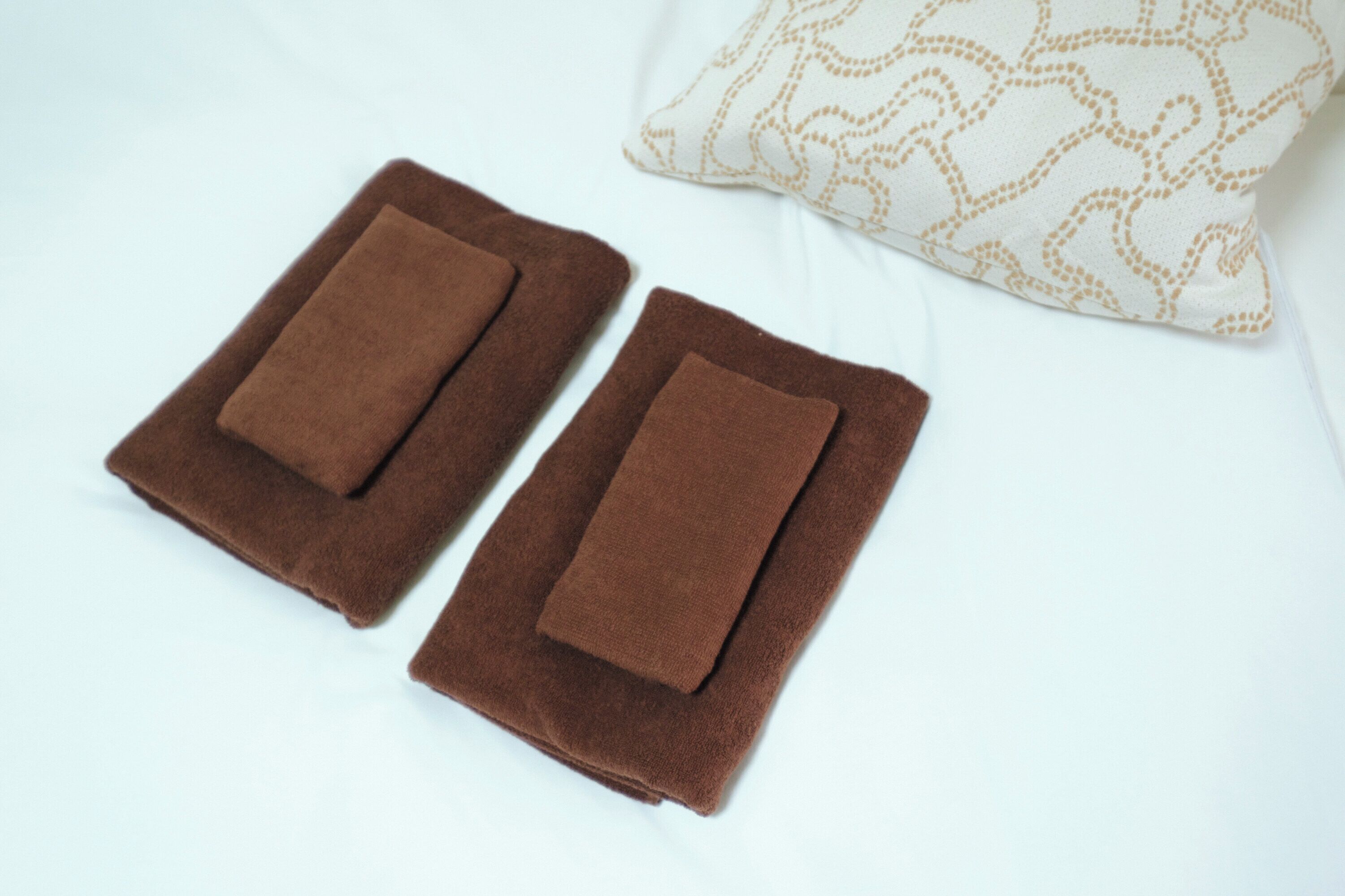 Two folded brown towels on a white bed with a patterned pillow.