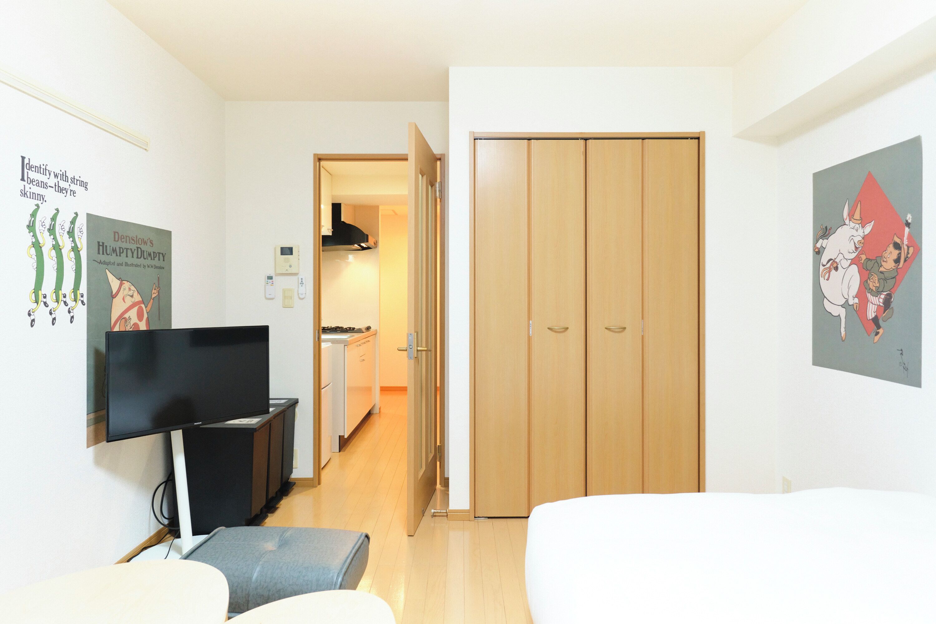 A hotel room with a TV, a sofa, a wooden wardrobe, and a kitchen in the background.