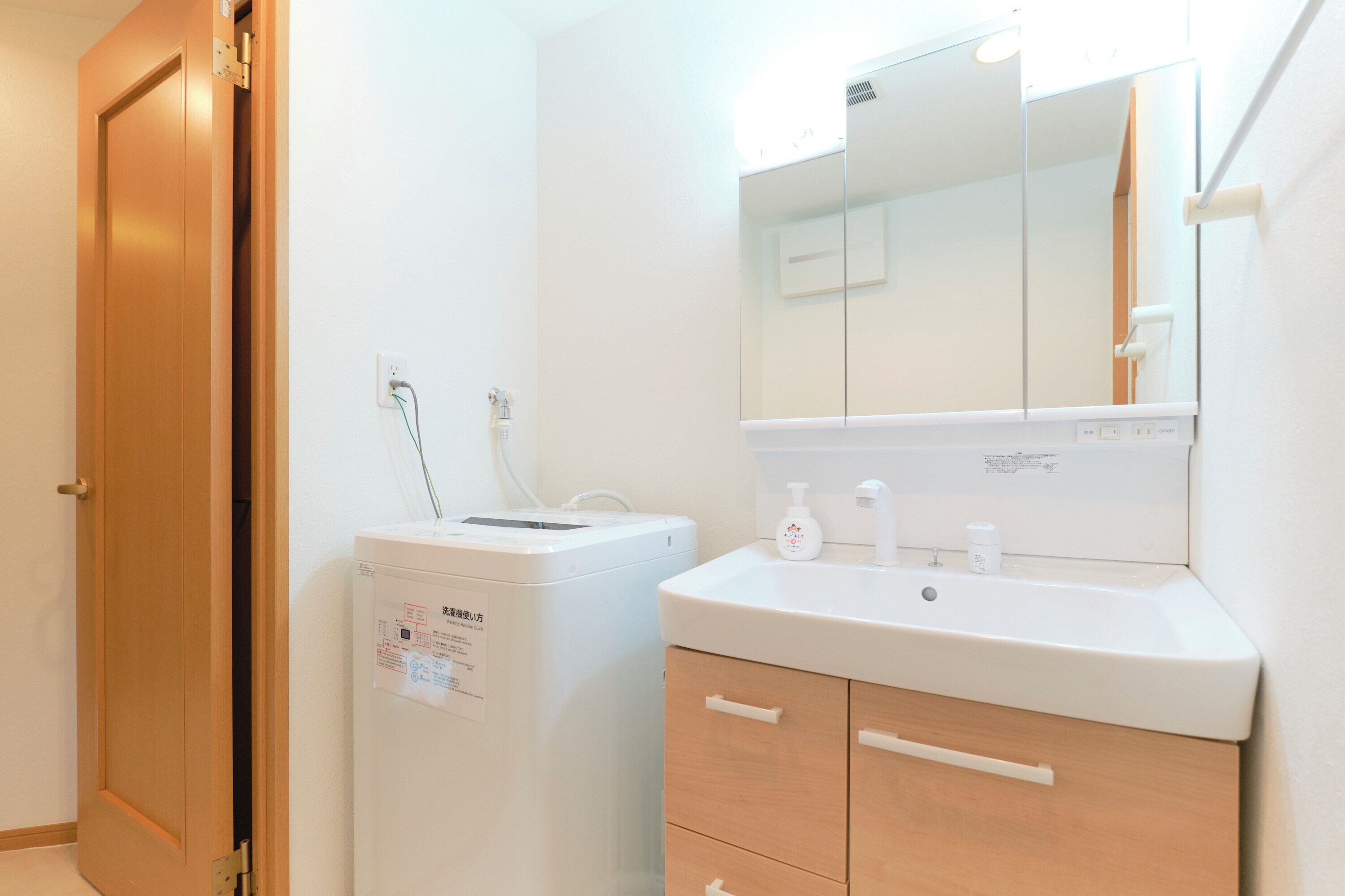 Une salle de bain avec une porte en bois, un lave-linge blanc, un lavabo avec un miroir et un meuble de rangement.