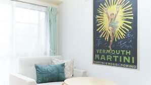 Una habitación con un sofá blanco, un cojín azul aguamarina y una mesa redonda. En la pared hay un cartel de Vermouth Martini.