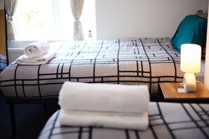 A neatly made bed with a patterned bedspread, a rolled towel on the bed, a bedside lamp, and a window with curtains. - Ol-mumps Cosy 2bd Free Parking & Wifi (Oldham)