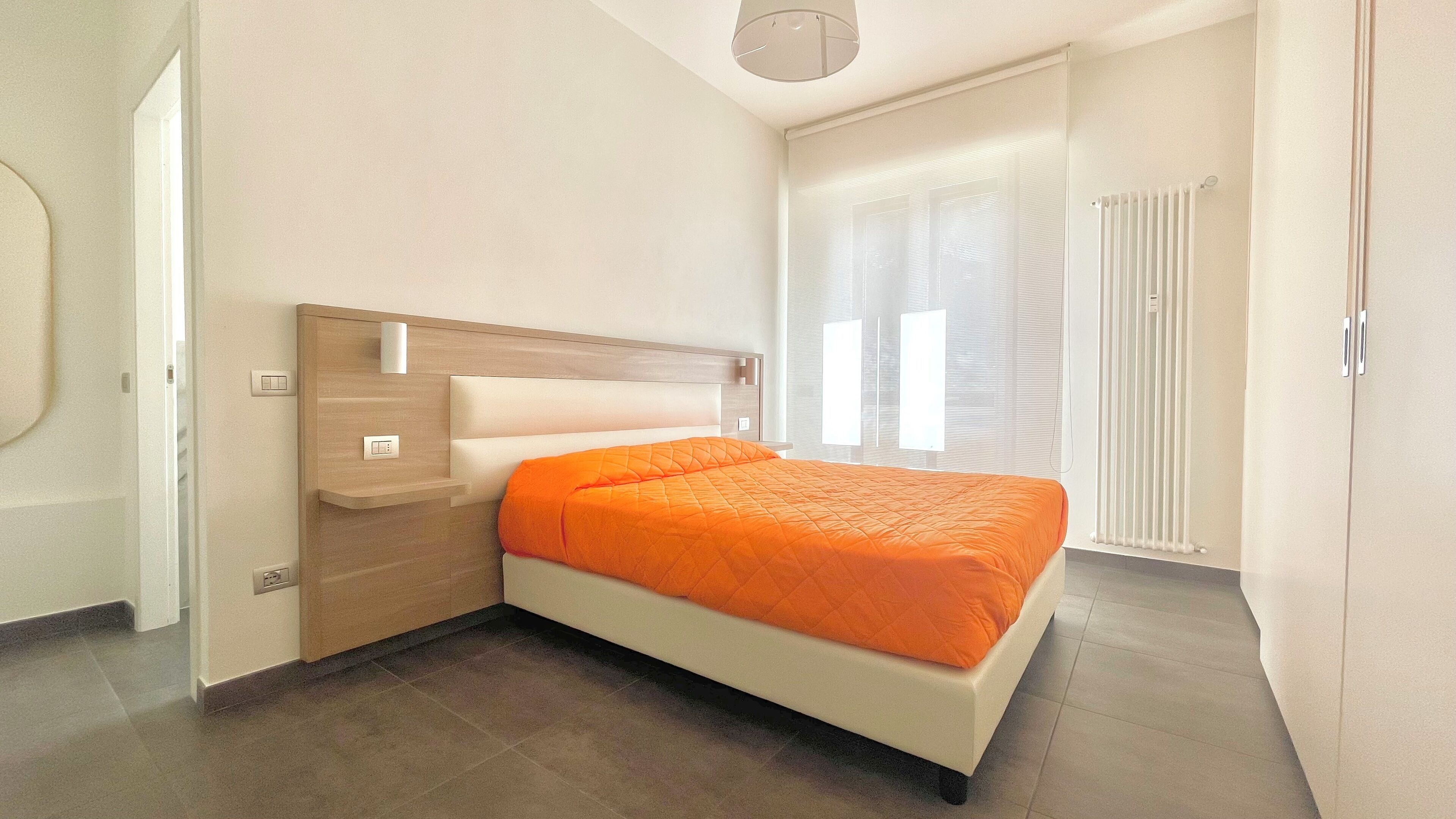 Quarto com cama, mesa de cabeceira, luminária na parede, janela com persianas e radiador.