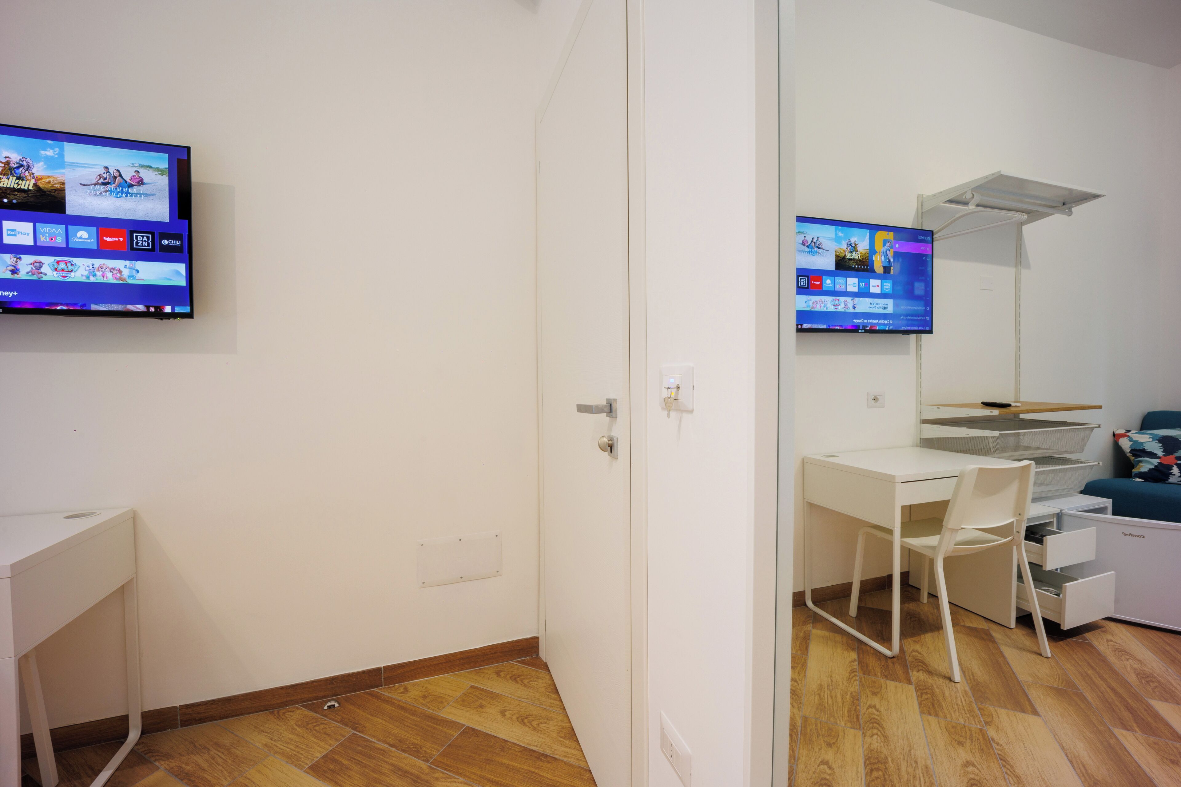 Una habitación moderna con un escritorio blanco, una silla, un televisor montado en la pared y un mueble auxiliar pequeño.