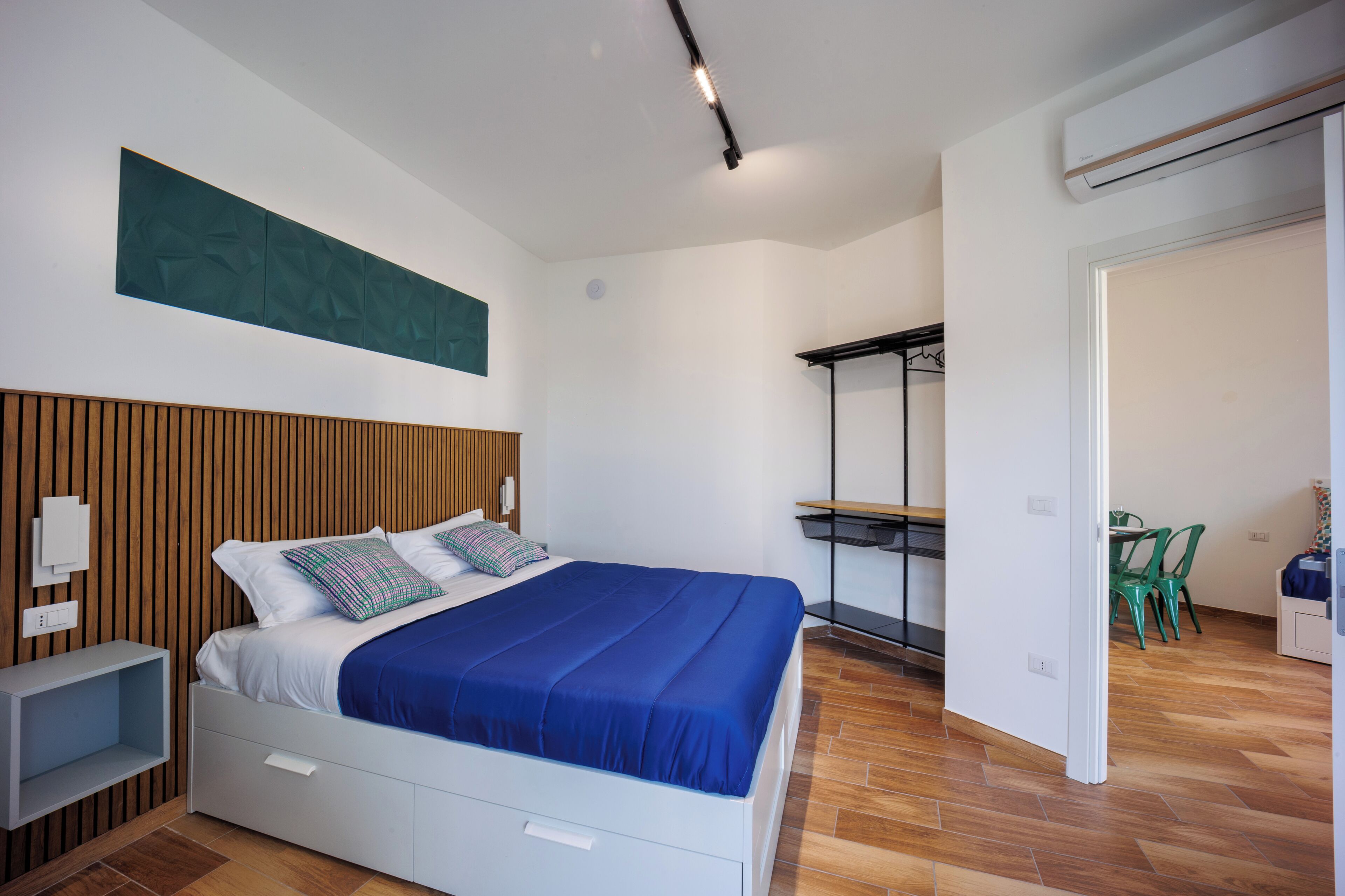 A modern bedroom with a wooden headboard, a large bed with blue bedding, a bedside table, and a small desk with a chair.