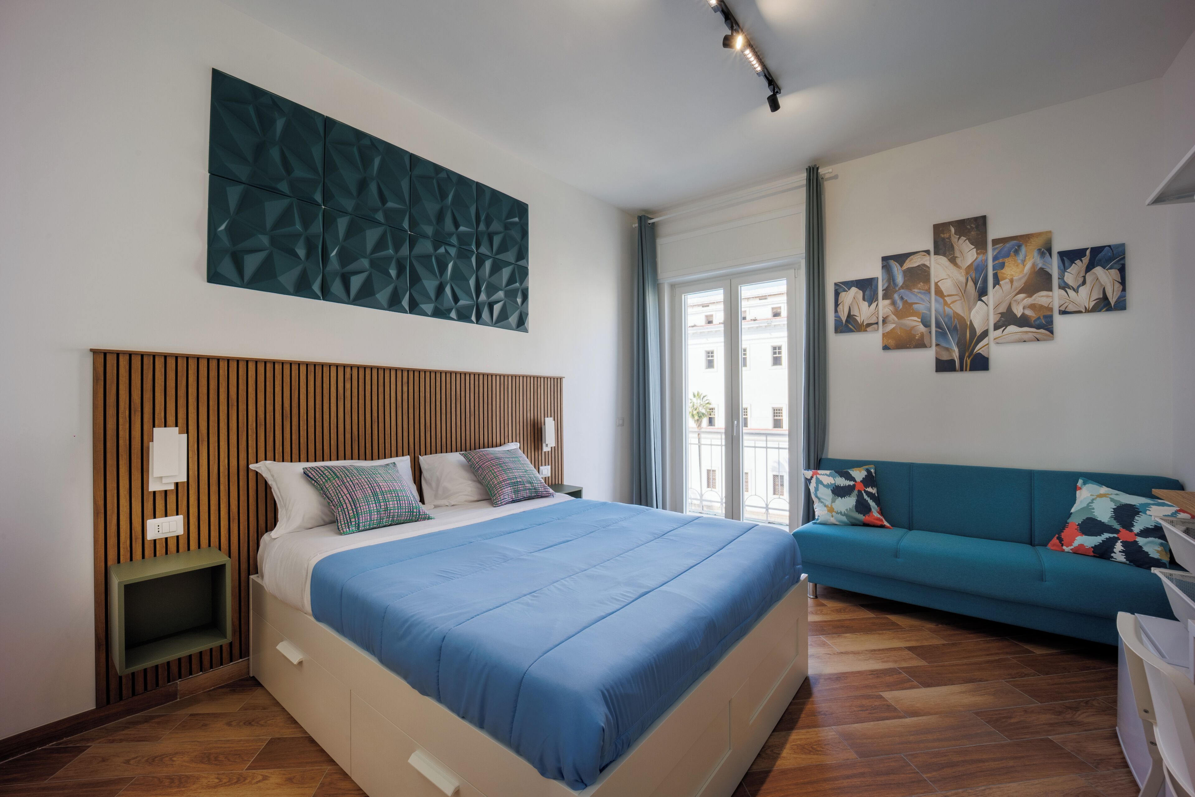 A modern bedroom with a wooden headboard, a large bed with blue bedding, a teal sofa, and a city view through the window.