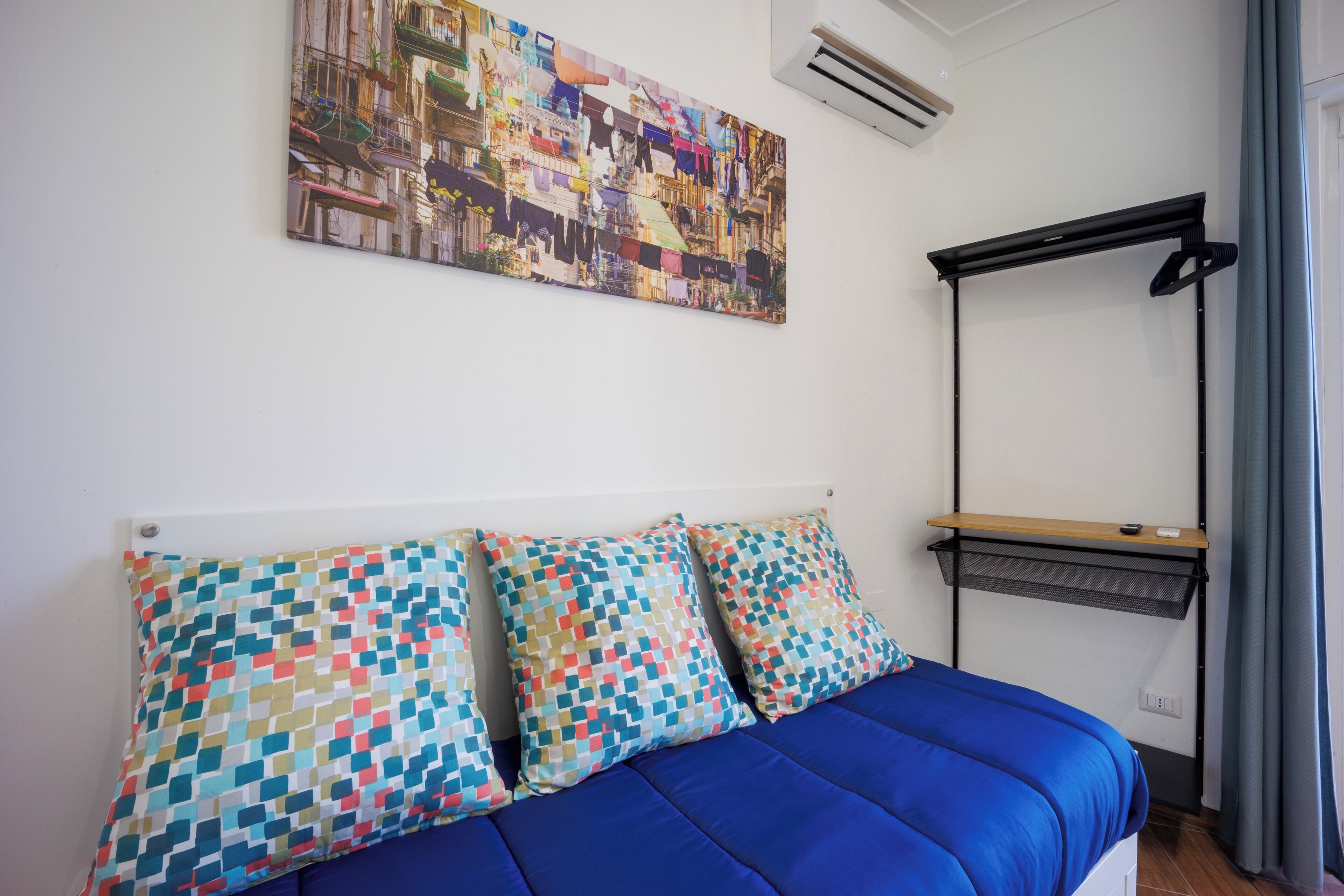 A bedroom with a bed, colorful pillows, a wall-mounted air conditioner, a window with curtains, and a hanging light fixture.