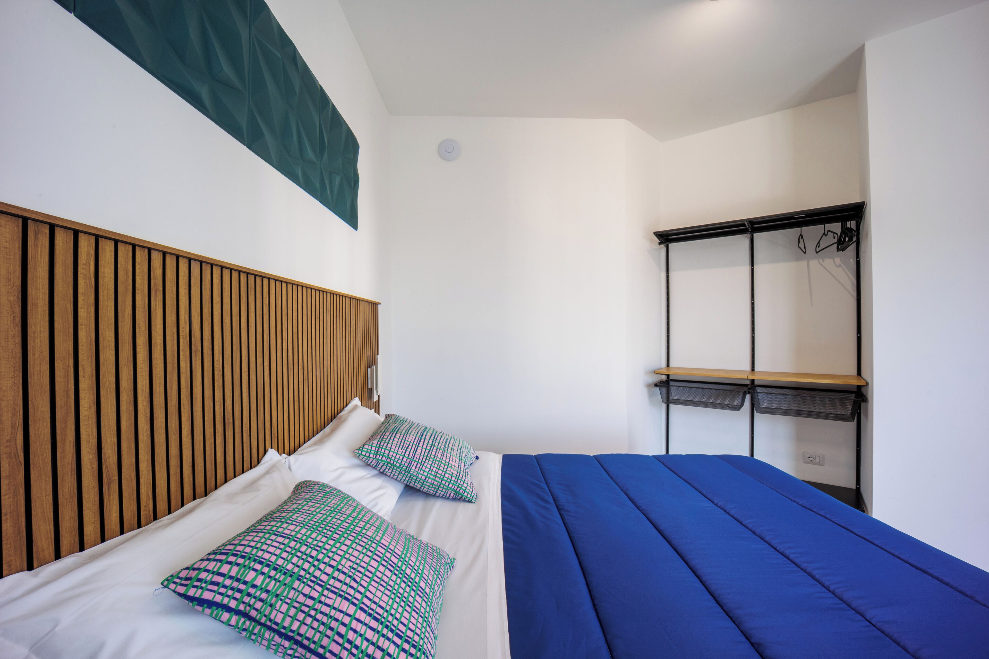A bedroom with a wooden headboard, a bed with blue bedding, and a shelf with hanging items.