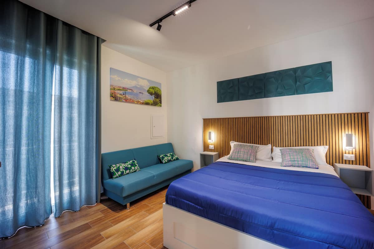 A bedroom with a wooden headboard, a blue bedspread, a teal sofa with patterned cushions, a painting on the wall, and a wooden floor.