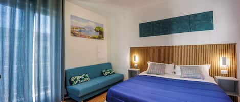 A bedroom with a wooden headboard, a blue bedspread, a teal sofa with patterned cushions, a painting on the wall, and a wooden floor.