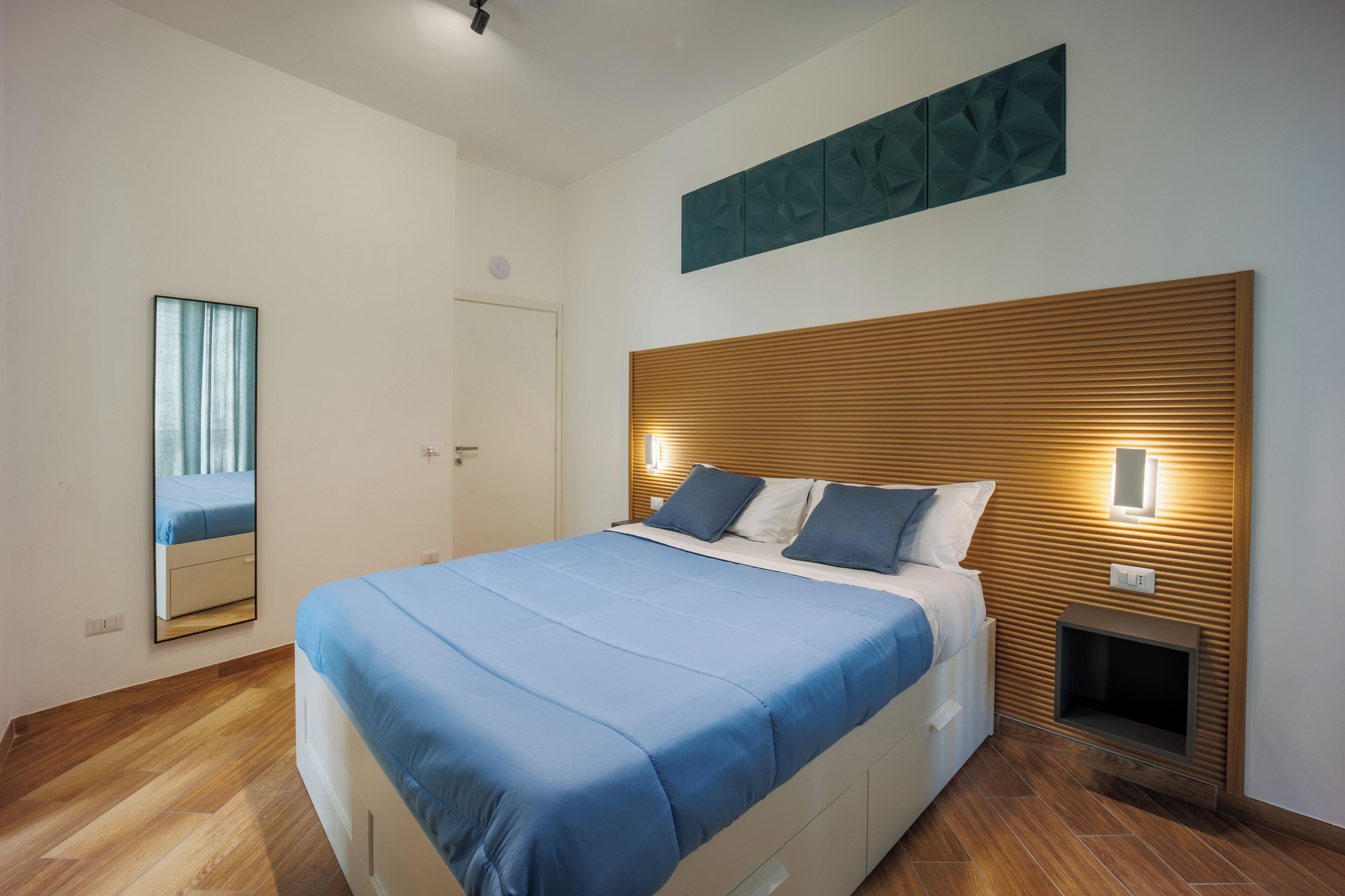 A modern bedroom with a large bed, wooden headboard, bedside lamps, and a mirror on the wall.