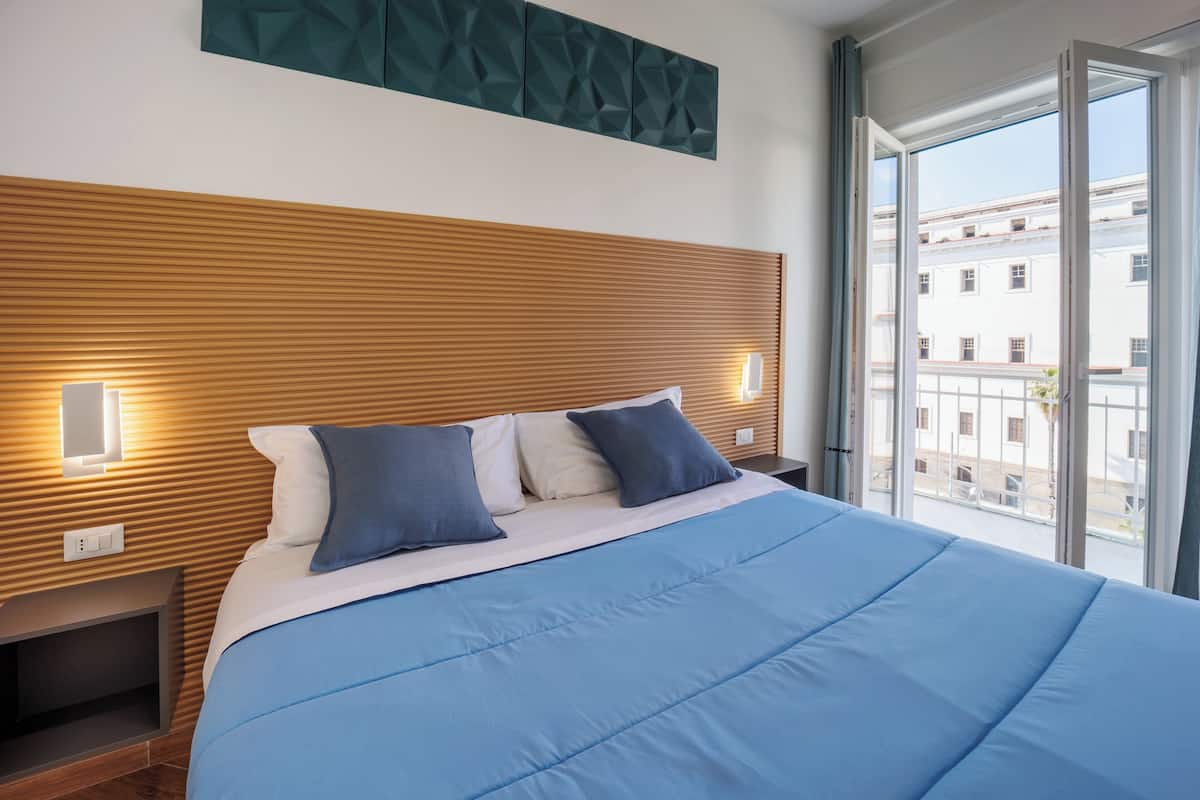 A modern hotel room with a wooden headboard, a large bed with blue bedding, and a view of buildings through the window.
